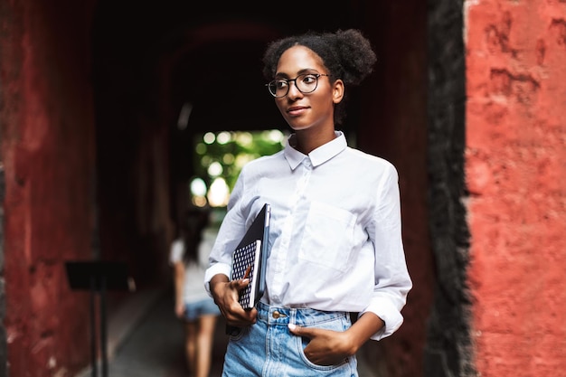 Bonita chica africana con anteojos y camisa sosteniendo una computadora portátil y un bloc de notas en las manos mientras mira cuidadosamente a un lado al aire libre