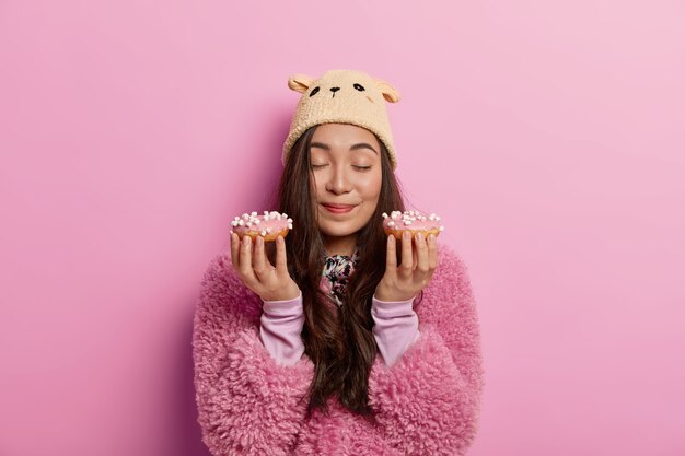 Bonita chica asiática disfruta de un agradable olor de deliciosas donas recién horneadas, tentadora por deliciosas donas, tiene un dilema de dieta, vestida con un abrigo rosa