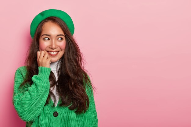 Bonita chica asiática hermosa con expresión alegre, viste boina verde de moda y jersey de punto, toca la barbilla, imagina un momento agradable en mente