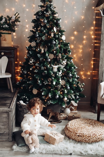 Bonita chica caucásica con el pelo rizado se sienta cerca de un gran árbol de Navidad hermoso