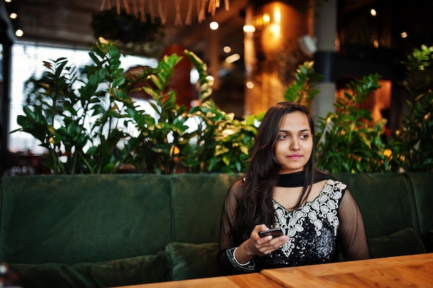Foto gratuita bonita chica india con vestido de sari negro posó en el restaurante con teléfono móvil a mano