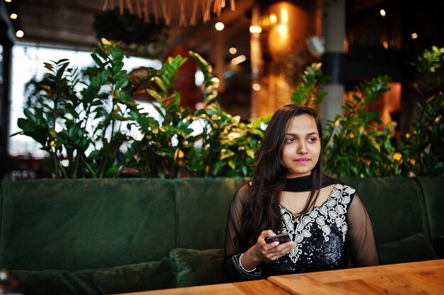 Bonita chica india con vestido de sari negro posó en el restaurante con teléfono móvil a mano