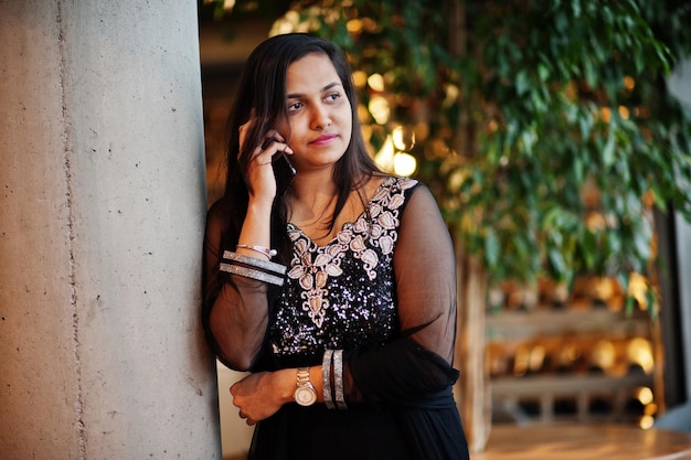 Bonita chica india con vestido de sari negro posó en el restaurante con teléfono móvil a mano