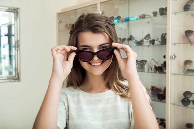 Bonita chica morena sonriendo, probándose gafas de sol en la tienda de óptica