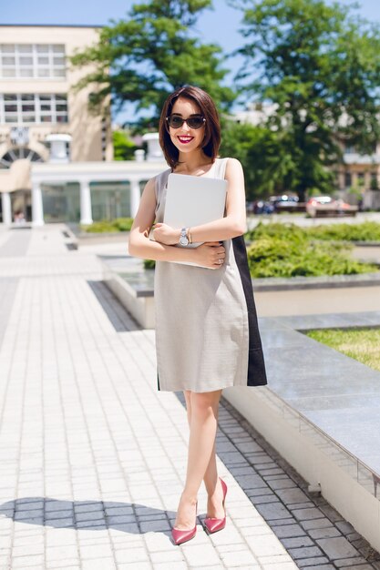 Bonita chica morena con vestido gris y tacones vinoso está de pie en el parque de la ciudad. Ella sostiene una computadora portátil y tiene una sonrisa amigable con lápiz labial vinoso.