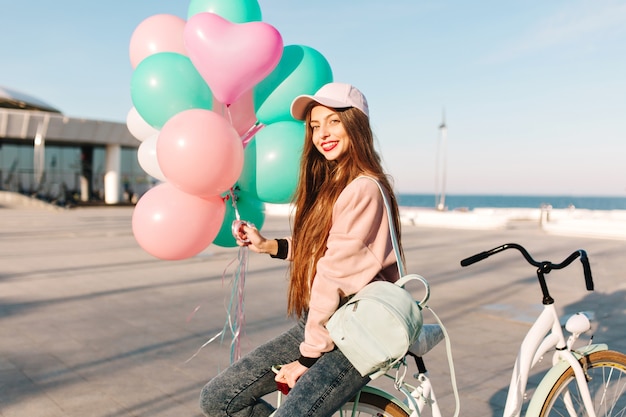 Bonita chica de pelo largo con traje rosa sentada en la bicicleta con globos esperando amigo del viaje.