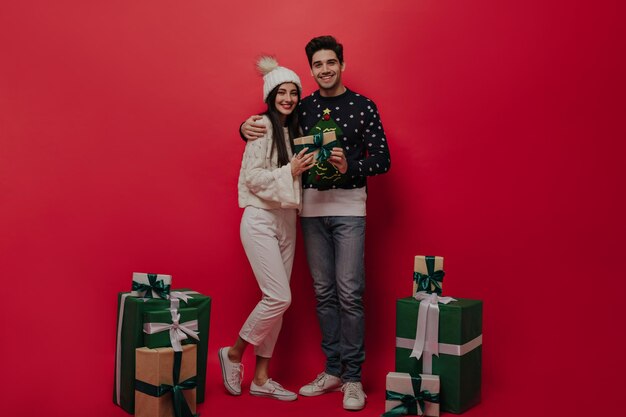 Bonita chica de pelo moreno con jersey de punto ligero, pantalones blancos, sombrero con pompón y hombre de pelo oscuro con lindos vaqueros y zapatillas sonriendo y posando cerca de cajas de regalo con fondo rojo.