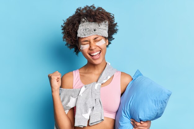 Bonita chica de pelo rizado llena de alegría aprieta el puño con alegría celebra finalmente alcanzar las metas descubre noticias positivas viste ropa de dormir tiene una expresión alegre sostiene la almohada aislada en la pared azul