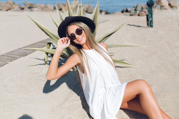 Bonita chica rubia con cabello largo está sentada en la playa cerca de cactus en el fondo. Ella está sosteniendo gafas de sol y sonriendo.