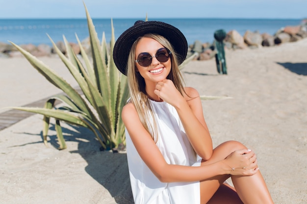 Bonita chica rubia con cabello largo está sentada en la playa cerca de cactus en el fondo. Ella sonríe a la cámara.