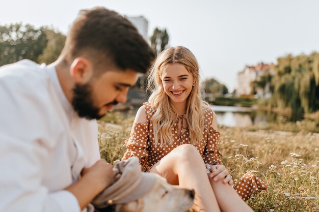 Bonita chica rubia y su amable marido descansan sobre la hierba y juegan con su mascota. Pareja posando con el telón de fondo del lago.