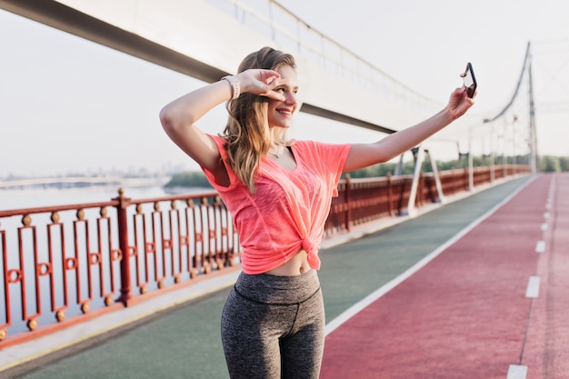 Bonita corredora con smartphone para selfie en pista de ceniza. Chica sensual en ropa deportiva tomando una foto de sí misma.