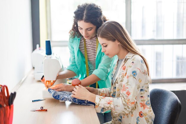 Bonita costurera enseñando a una chica que trabaja con una máquina de coser en clases de costura con una ventana en el fondo en un taller de costura moderno