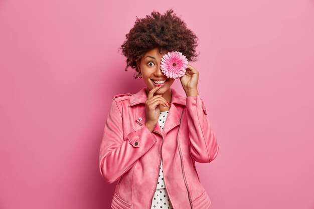 Bonita dama de cabello rizado con una sonrisa dentuda mantiene una flor de gerbera rosa frente a los ojos, flores recogidas de los prados florecientes de primavera, vestida con una chaqueta rosa, va a hacer una corona