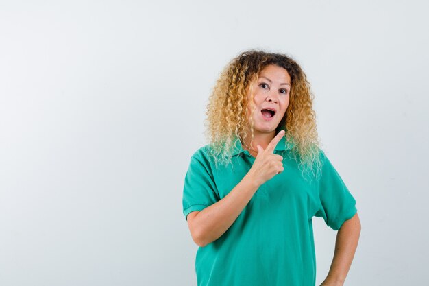 Bonita dama rubia apuntando a la esquina superior derecha con una camiseta de polo verde y mirando asombrada. vista frontal.