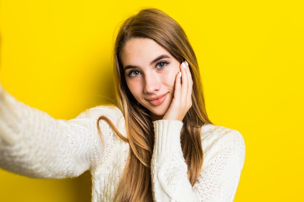 Bonita encantadora sonriente chica de moda soleada está haciendo selfie en su teléfono con un suéter blanco ancho en amarillo