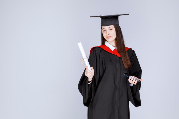 Bonita estudiante de posgrado en bata de teléfono móvil en mano sosteniendo diploma. Foto de alta calidad