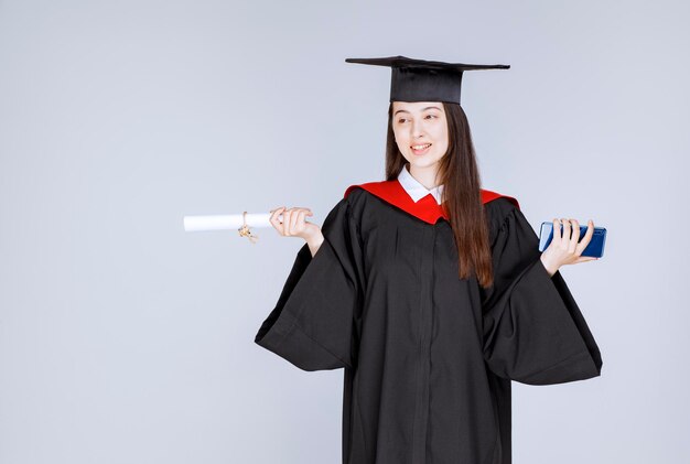 Bonita estudiante de posgrado en bata de teléfono móvil en mano sosteniendo diploma. Foto de alta calidad