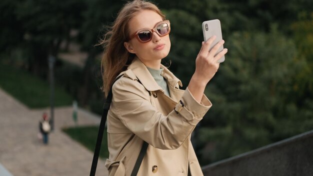 Bonita estudiante vestida con una gabardina con gafas de sol tomando selfie en un smartphone con una apariencia segura al aire libre Tecnología moderna