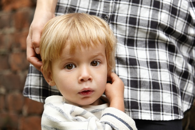 Bonita foto de niño rubio inocente con ojos marrones. Hermosa expresión facial infantil con la boca ligeramente abierta. Bebé de pie junto a la joven madre en camisa a cuadros. Mamá acariciando la cabeza del niño cerca de la pared de ladrillo.