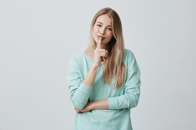 bonita hembra mantiene el dedo en los labios sonrientes, muestra un signo de silencio, pide mantener la conspiración o el silencio, trata de ocultar el secreto