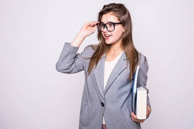 Bonita y joven estudiante con gafas grandes cerca de algunos libros sonriendo sobre fondo blanco.