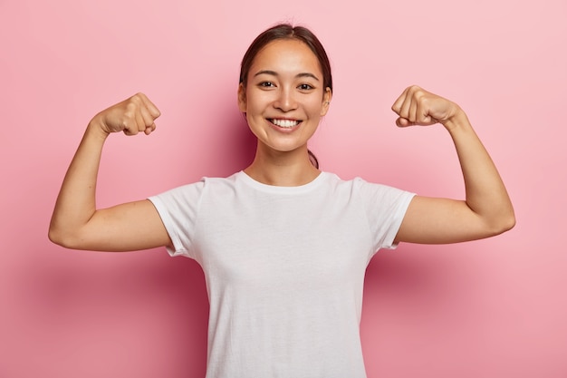 Bonita modelo femenina coreana se mantiene en forma y saludable, levanta las manos y muestra los músculos, se siente orgullosa de sus logros en el gimnasio, sonríe ampliamente, se viste con ropa casual blanca, posa en interiores muestra un poder real