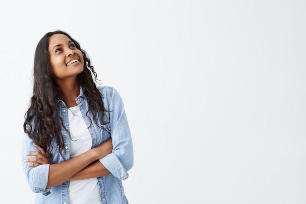 Bonita morena afroamericana con cabello largo y ondulado mirando hacia arriba y lejos, sonriendo sinceramente, manteniendo los brazos cruzados sobre la camisa vaquera. Concepto de personas y emociones positivas.