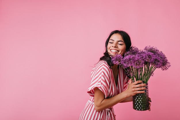 Bonita morena está sonriendo deslumbrantemente mientras disfruta de las flores presentadas. Retrato de niña en top rayado rosa cerrando los ojos de felicidad.