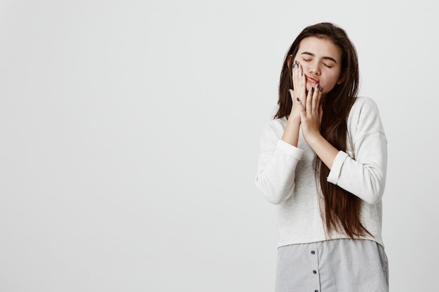 Bonita morena hermosa mujer adolescente con su largo cabello lacio y oscuro suelto en ropa casual tocando las mejillas con las manos, posando contra la pared gris con los ojos cerrados.