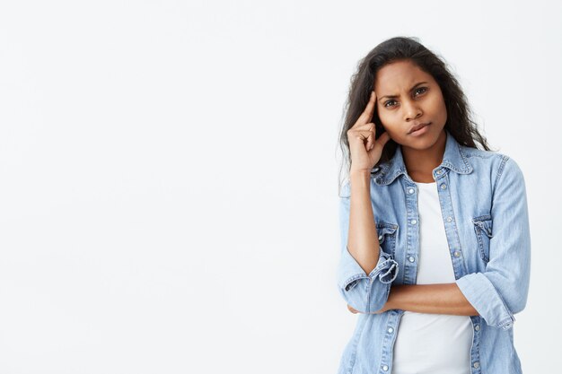 Bonita morena mujer afroamericana con el pelo largo y ondulado frunciendo el ceño mientras que con mirada plácida y pensativa. Pensativa mujer de piel oscura con expresión perpleja pensando