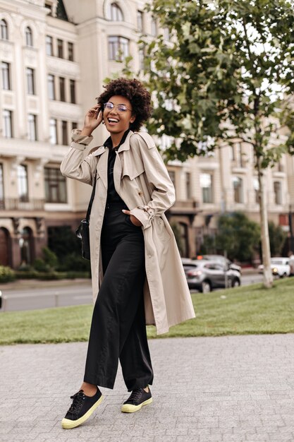 Bonita morena rizada de buen humor en pantalones negros de gran tamaño, gabardina beige y anteojos sonríe y posa afuera