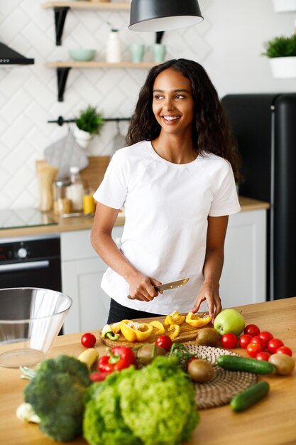 Bonita mujer afro corta un pimiento amarillo y sonriendo mira en la ventana