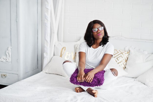 Bonita mujer afroamericana con anteojos posada en la habitación sentada en la cama con almohada violeta