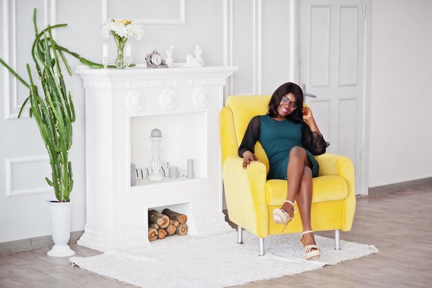 Bonita mujer afroamericana con anteojos posada en la habitación sentada en una silla amarilla