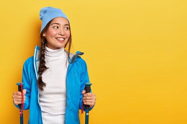 Bonita mujer asiática sonriente disfruta de caminar escandinavo, tiene un viaje de senderismo, mira a un lado, tiene trenza peinada, usa sombrero azul y chaqueta, sostiene bastones de trekking, aislado en una pared amarilla