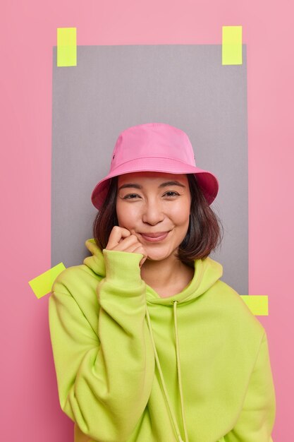 Bonita mujer asiática tiene una mirada suave a la cámara, mantiene la mano en la mejilla, usa panamá y una sudadera con capucha verde se siente feliz posa contra el espacio de copia en blanco para su contenido publicitario
