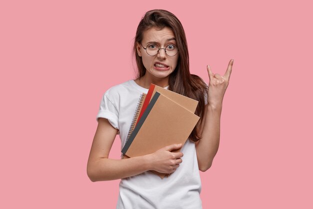 Bonita mujer con cabello oscuro, hace gestos de rock n roll, se siente genial, lleva blocs de notas, vestida con una camiseta blanca informal, modelos sobre espacio rosa