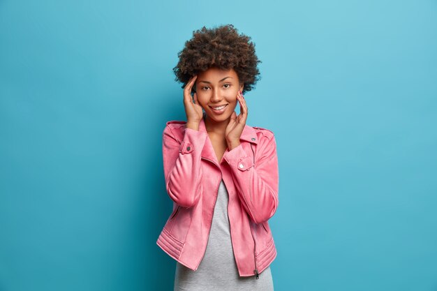 Bonita mujer encantadora con tierna expresión, sonríe agradablemente, toca la cara con ternura, se ve confiada, vestida con ropa de moda,