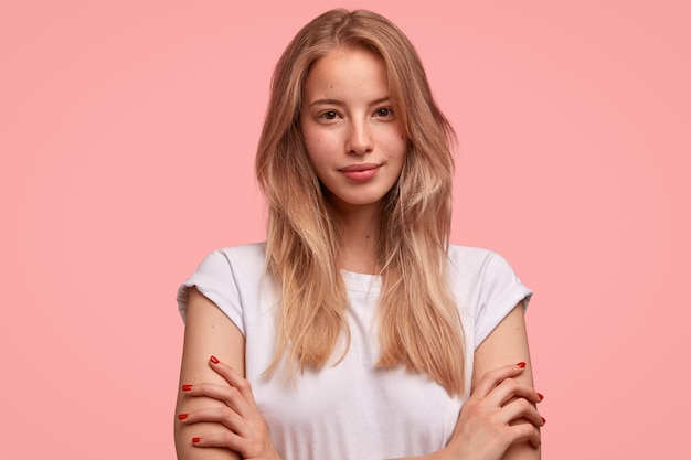 Bonita mujer europea con cabello lacio, mantiene las manos cruzadas, viste una camiseta blanca informal, se para contra la pared rosa