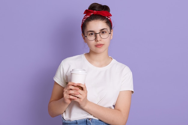 Bonita mujer europea morena tiene cabello oscuro y nudo, viste camiseta blanca casual y banda de aire roja, sostiene café para llevar