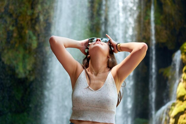 Bonita mujer con gafas de sol jugando con su cabello mientras está de pie cerca de una cascada