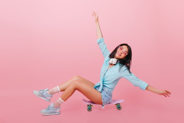 Bonita mujer hispana con piel bronceada agitando las manos mientras está sentado en longboard. Chica latina inspirada en gafas de sol posando en patineta