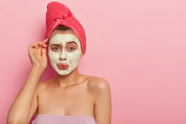 Bonita mujer joven con expresión avergonzada, se aplica mascarilla de crema en la cara para reducir los problemas de la piel, tiene mirada de descontento, se baña todos los días, disfruta de los procedimientos higiénicos. Cuidado de la salud