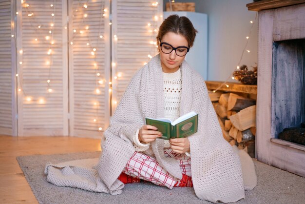 Una bonita mujer joven con gafas está sentada junto a la chimenea en una manta leyendo un libro