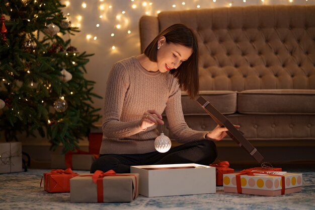 Bonita mujer morena abre un adorable regalo sosteniendo un globo de Navidad blanco