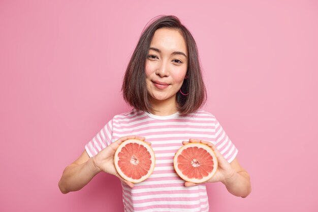 Bonita mujer morena con cabello oscuro sostiene dos mitades de pomelo sobre el cuerpo vestido con una camiseta a rayas casual que se ve satisfecha aislada sobre una pared rosa. La hembra sostiene la fruta jugosa