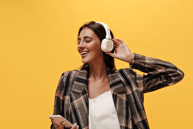 Bonita mujer morena con camiseta blanca y chaqueta marrón sonríe ampliamente y escucha música en los auriculares Linda chica sostiene el teléfono sobre fondo amarillo