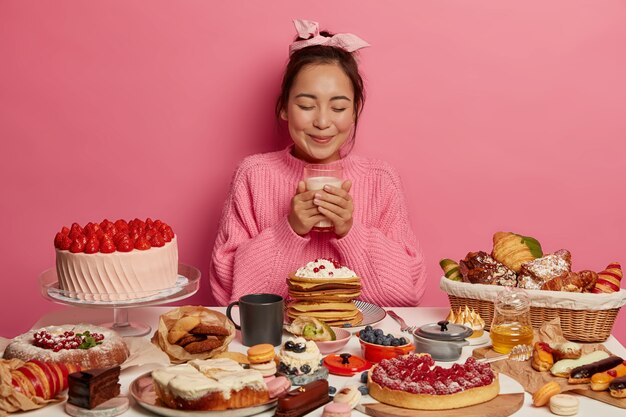 Bonita mujer morena sostiene un vaso de leche, come confitería y dulces, usa un suéter de punto y una diadema, siendo golosa posa en la mesa festiva contra el fondo rosa.