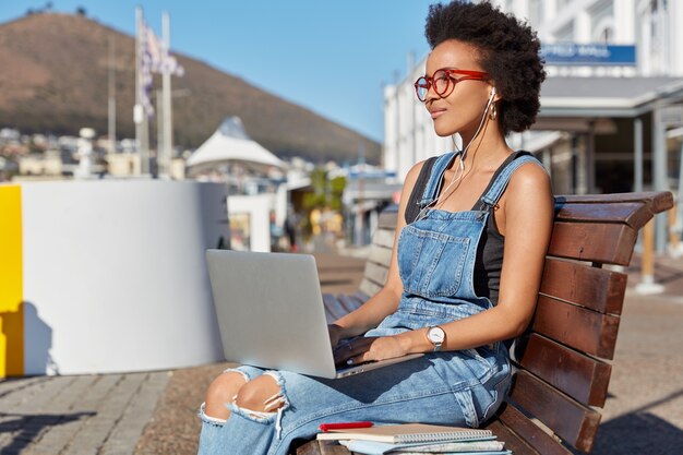 Bonita mujer negra mira el espectáculo con auriculares y portátil, disfruta de un volumen alto, escucha audiolibros, se prepara para las clases, da un paseo durante el día soleado de verano, usa overoles de mezclilla, navega por Internet